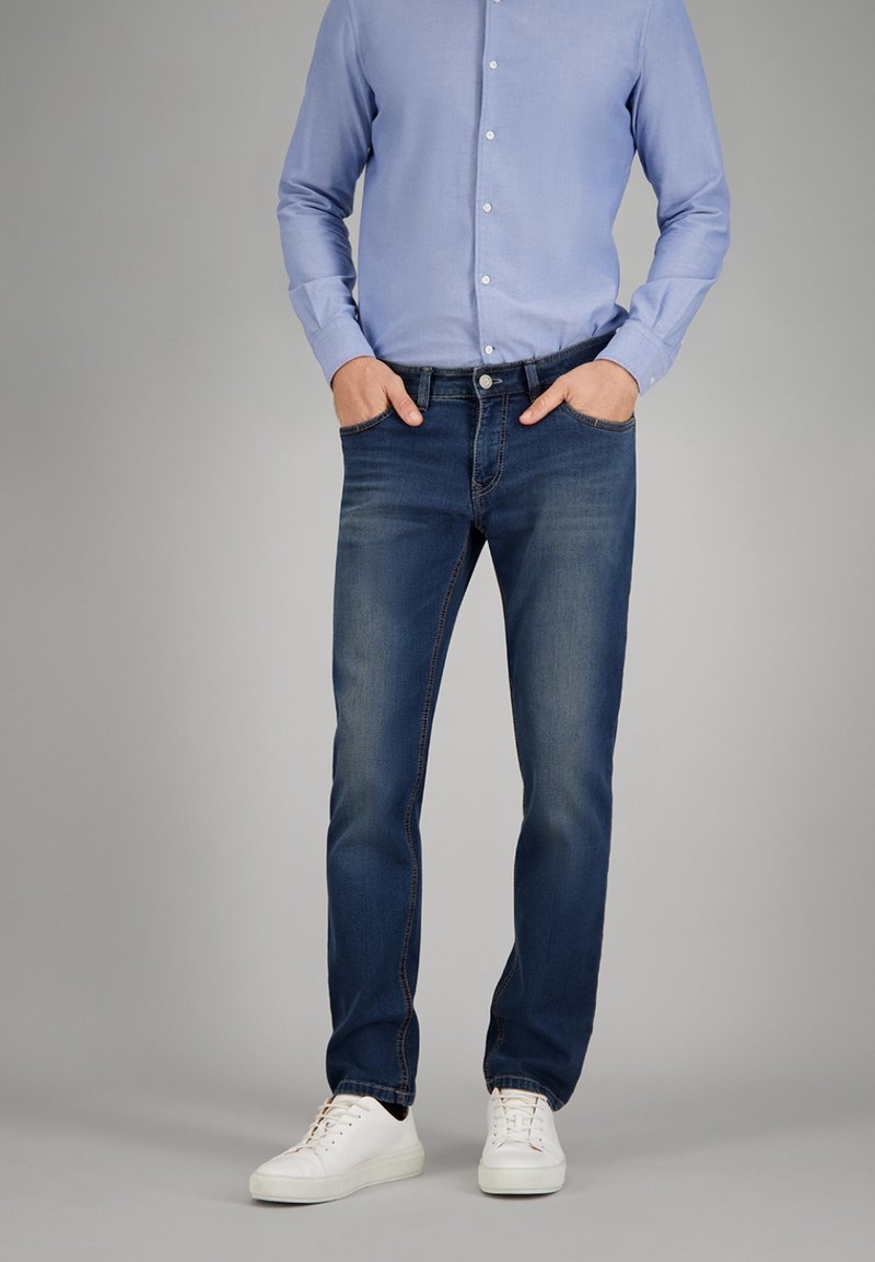 Donkerblauwe denim jeans met een rechte pasvorm, voorzien van een klassiek vijf-pocket ontwerp, zichtbare stiksels en versleten details, gecombineerd met een lichtblauw shirt en witte sneakers.