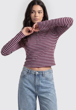 Kvinde med langt mørkt hår iført en vinrød og grå stribet langærmet crop top og blå jeans, der poserer med armene hævet nær ansigtet.