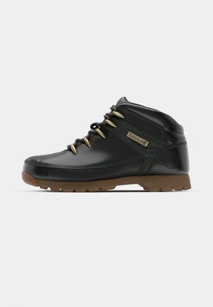 Black leather ankle boots with a textured green panel, gold eyelets, and a brown rubber sole. Features Timberland logo on the side.