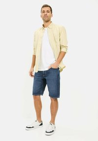 Lys beige skjorte med knapper over en hvid T-shirt, parret med blå denim shorts og hvide sneakers med mørke accenter.