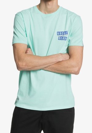 Homme portant un T-shirt turquoise clair avec le texte « VACAY SUMMER BEACH » sur la poitrine, bras croisés, associé à un pantalon noir.
