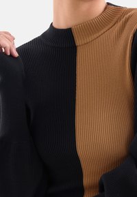 Zbliżenie na sweter z prążkowanej dzianiny w czarno-brązowe pionowe pasy i wysoki dekolt noszony przez osobę.