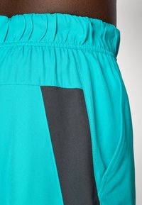 Shorts de sport turquoise avec une taille élastique, un panneau latéral gris contrasté et une texture de tissu lisse et léger.