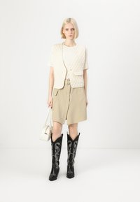 Quiltet elfenben vest, ribbet krem topp, beige skjørt med lommer, og svarte broderte cowboystøvler. Inkluderer en liten hvit håndveske.