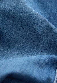 Gros plan sur un tissu en denim bleu texturé avec un motif de tissage visible et de délicats plis créant des ombres douces.