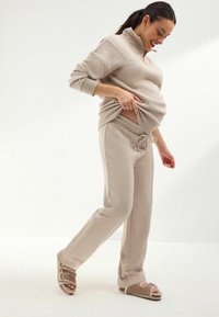 Beige Kapuzenpullover mit Reißverschluss und passender Jogginghose aus weichem Material, mit einem Kordelzug an der Taille. Getragen mit braunen Sandalen mit Schnallen.