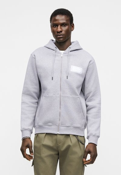Άνδρας που φοράει ανοιχτό γκρι φούτερ με φερμουάρ, εμπρός τσέπες και κορδόνια, σε συνδυασμό με χακί παντελόνι και ασημένιο βραχιόλι στον αριστερό καρπό.