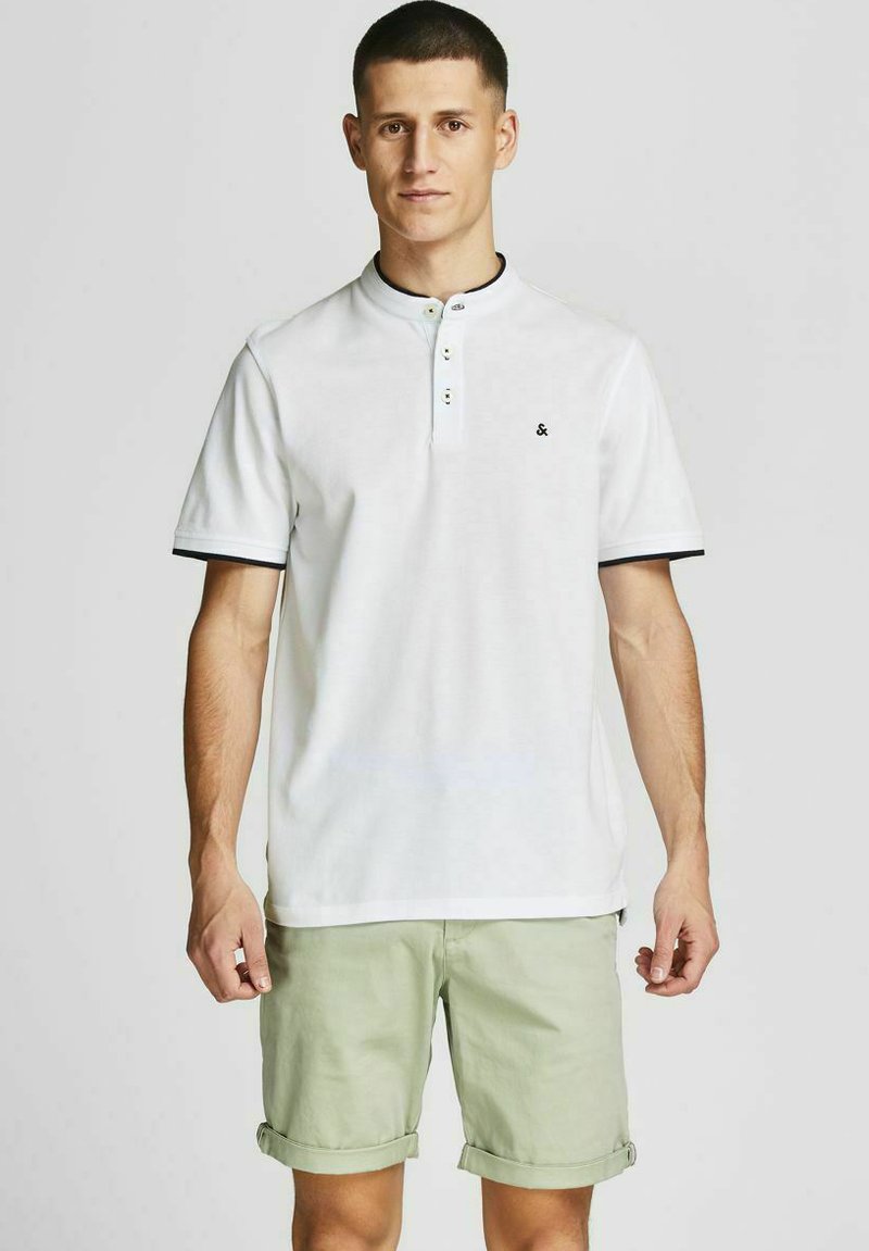 Witte poloshirt met een twee-knopenknopsluiting, zwarte kraagafwerking en klein logo op de borst. Gepaard met lichtgroene opgerolde shorts.