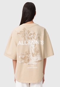 Beżowa, oversize'owa koszulka z bawełny z nadrukiem anioła w delikatnej bieli, z napisem "ALLSAINTS LONDON" i "THE SAINTHOOD" poniżej.