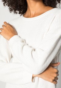 Vit stickad tröja med ett strukturerat mönster, ribbad mudd och rund halsringning, som visar ett mjukt och andningsbart tyg.