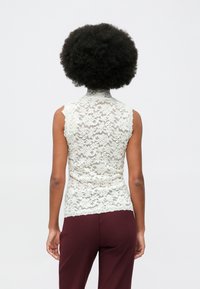 Person med krøllete afrofrisyre iført en ermeløs hvit blondetopp og marsalrøde bukser, vendt bort fra kameraet mot en ensfarget bakgrunn.