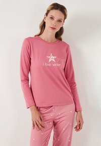 Top de manga larga rosa con el texto en blanco "Siempre amo el invierno" y un gráfico de estrella, combinado con pantalones rosas que tienen un patrón de estrellas blancas. Tejido suave.