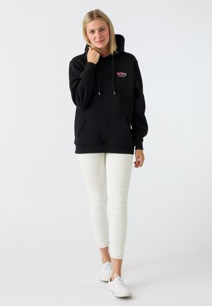 Schwarzer Kapuzenpullover mit einer Fronttasche, versehen mit einem rosa Herzdesign auf der Brust. Kombiniert mit weißen Slim-Fit-Hosen und weißen Sneakern.