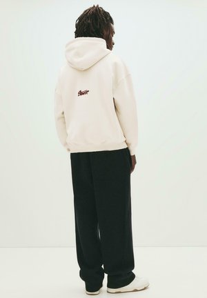 Persona con dreadlocks indossa un ampio hoodie beige con logo sul retro, pantaloni larghi neri e sneakers bianche, in piedi contro uno sfondo chiaro e semplice.