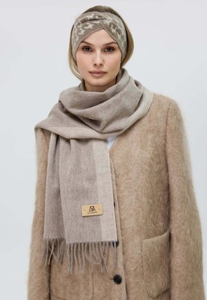 Light beige scarf made of soft, textured fabric, featuring fringed ends and a small logo tag. Worn with a fluffy light brown coat.