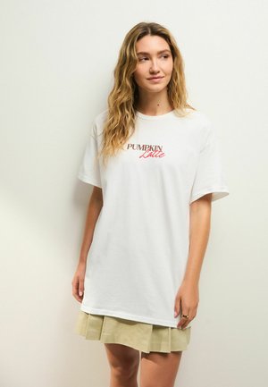 T-shirt bianco oversize in cotone, con la scritta "PUMPKIN Latte" stampata in testo marrone e rosso. Indossato con una gonna plissettata beige chiaro.