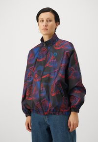 Windbreaker jacket in black with red and blue rose patterns, zip closure, high collar, and elastic cuffs. Features logo patch on the chest.