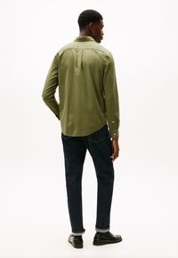 Groene blouse met lange mouwen en een button-down kraag, met plooidetail op de rug, gecombineerd met donkerblauwe jeans en zwarte schoenen, gezien van achteren.