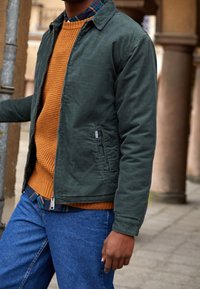 Groene canvasjas over een oranje structuurtrui, met daaronder een blauw geruit overhemd, gecombineerd met blauwe spijkerbroek.