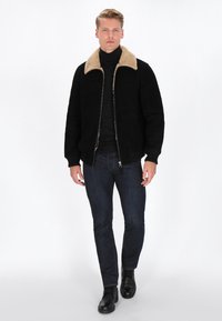 Schwarze Wildlederjacke mit beigem Lammfellkragen, Reißverschluss vorne und gerippten Bündchen, getragen über einem schwarzen Rollkragenpullover und blauen Jeans.
