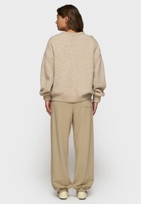 Mujer con cabello castaño hasta los hombros de espaldas, vestida con un suéter oversized beige, pantalones beige holgados y zapatos color crema.