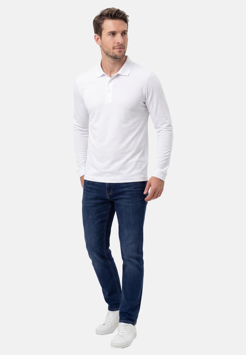 Homme portant un polo à manches longues blanc, un jean bleu foncé et des baskets blanches, debout les mains dans les poches.