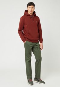 Bordeaux hoodie met trekkoord, voorzak en groene broek. Grijze sneakers met rode accenten. Eenvoudige achtergrond.