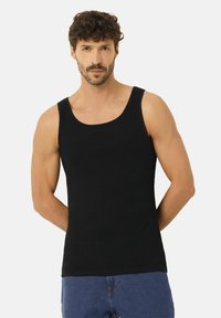 Mann mit kurzen lockigen Haaren und Bart, der ein schwarzes ärmelloses Tanktop und blaue Jeans trägt und mit den Händen hinter dem Rücken steht.