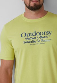 Limegroene katoenen t-shirt met korte mouwen, voorzien van donkerblauwe tekst die zegt "Buitengevoelens Vooruit! Abonneer op de Natuur®."