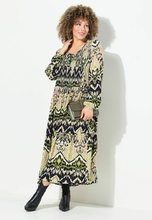 Maxi-jurk met een zwart, groen en roze tribale print; lange mouwen, een geplooide taille en een vloeiende silhouet. Gecombineerd met een groene clutch.