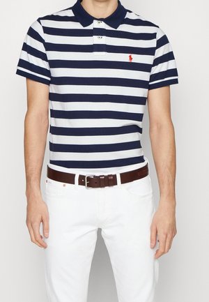 Poloshirt met marineblauwe en witte horizontale strepen, kraag met twee knopen en klein rood logo, gecombineerd met witte denim en een bruine riem.