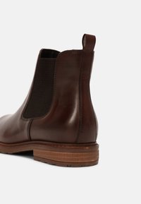 Botte Chelsea en cuir marron avec panneau latéral en élastique, languette de tirage au talon et semelle en bois superposée sur fond blanc.