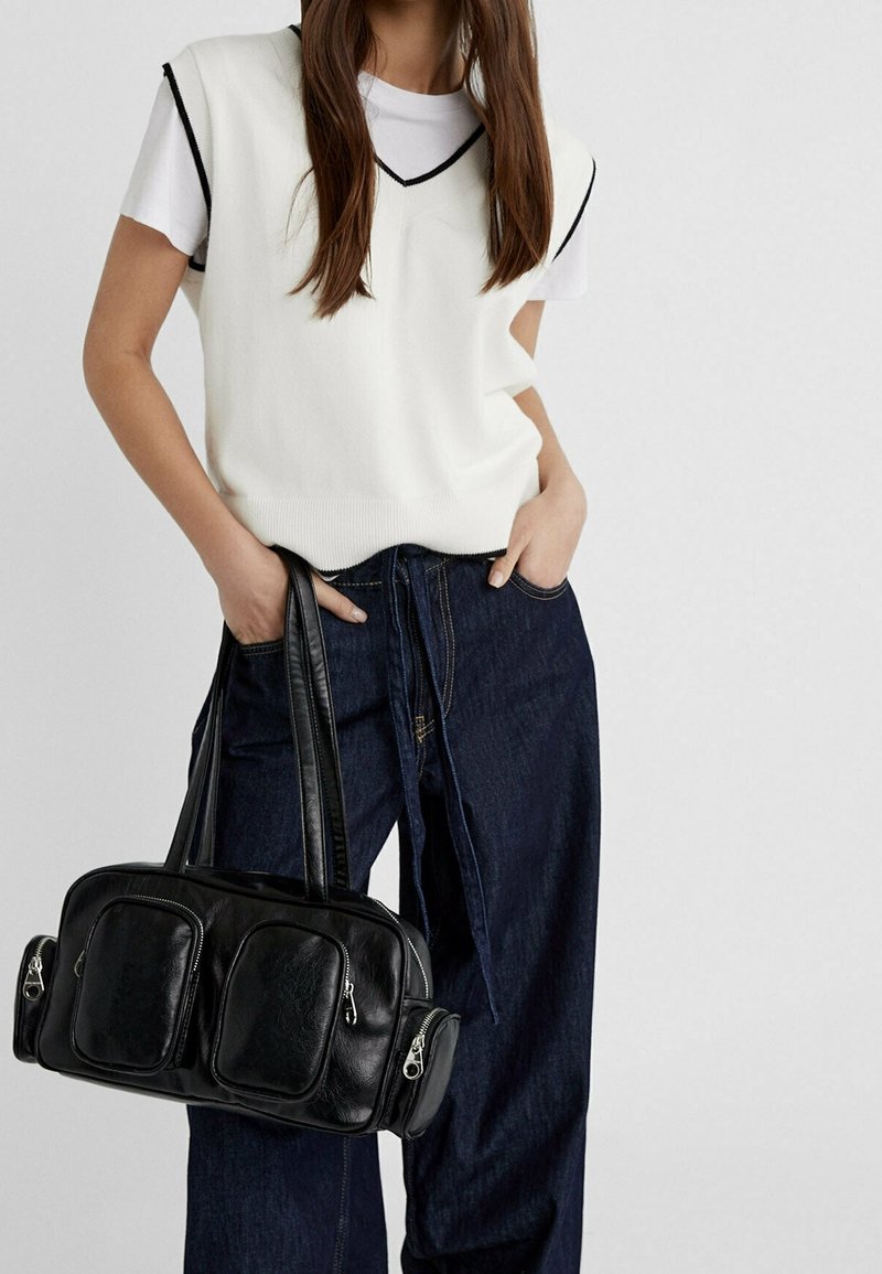 Femme portant un pull sans manches blanc avec bordure noire par-dessus un t-shirt blanc, jean large foncé, tenant un sac à main en cuir noir avec plusieurs poches.