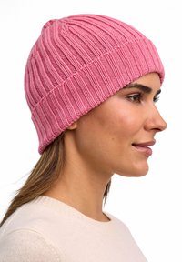Donna in un maglione crema che indossa un berretto rosa a coste, rivolta verso destra con un'espressione neutra su uno sfondo bianco.