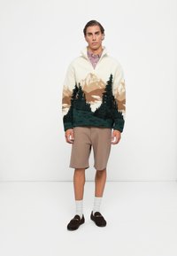 Sweater with a cream base featuring green and brown mountain landscape. Paired with beige shorts. Brown loafers and white socks complete the outfit.