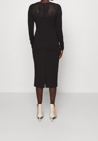 Robe noire à manches longues avec une coupe ajustée, longueur au genou, dotée d'un panneau arrière transparent et d'une fermeture éclair au dos. Portée avec des bottines beiges.