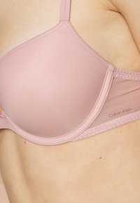Svijetloružičasti podstavljeni grudnjak s glatkom tkaninom, podešivim trakama i suptilnim detaljima šivanja. Ime branda "Calvin Klein" vidljivo je sa strane.