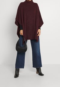 Poncho bordeaux con collo alto, realizzato in un morbido tessuto a maglia; abbinato a jeans a gamba larga blu scuro e stivaletti neri con tacco.