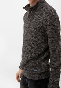 Stickad tröja i mörkgrått med hög krage, tvåknappsstängning, strukturerat mönster och en etikett på vänster sida nära fållen.