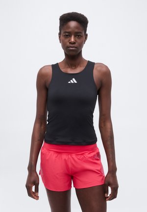 Atletisk kvinde iført en sort ærmeløs top med hvidt logo og lyse koralfarvede sportsshorts, stående mod en ensfarvet lys baggrund.