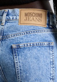 Šviesiai mėlynos džinsinės kelnės su odiniu etikete, ant kurios parašyta "MO5CH1NO JEANS". Su siuvinėjimu ir dviem galiniais kišenėmis. Bendras išblukęs vaizdas.