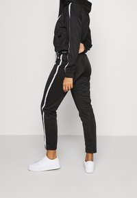 Svart träningsdräkt med en cropped jacka och slim-fit byxor, med vita sidostycken, elastiska ärmslut och vita sneakers.