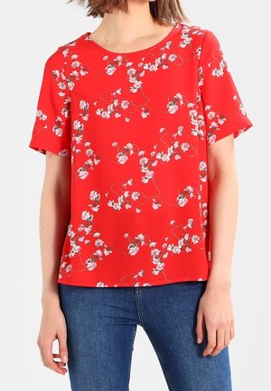 Vrouw die een rood shirt met korte mouwen en een wit bloemenpatroon draagt, gecombineerd met een blauwe spijkerbroek, staand tegen een effen achtergrond.