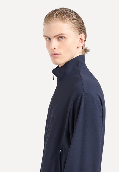 Giacca a zip blu navy con colletto alto, caratterizzata da una texture a costine e dettagli di cucitura discreti; progettata con una vestibilità slim e tasche laterali.