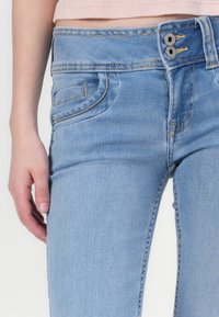 Ανοιχτό μπλε τζιν παντελόνι με στενή γραμμή και κλείσιμο με δύο κουμπιά, διαθέτοντας μπροστινές τσέπες και αντίθετη ραφή κατά μήκος των ραφών.