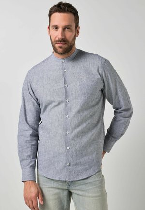 Hombre con barba, vestido con una camisa de cuello mandarín gris a rayas y jeans de lavado claro, de pie con una mano detrás de la espalda.