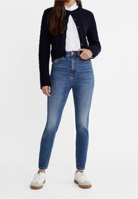 Cárdigan de punto de cable en navy sobre una camisa blanca de botones, combinado con jeans ajustados de talle alto en azul y zapatillas blancas con detalles en marrón.