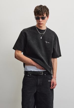 Jeune homme portant un t-shirt noir texturé oversize, un jean noir, des colliers argentés superposés, des lunettes de soleil, la main rentrée dans le pantalon.