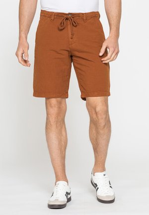 Uomo che indossa pantaloncini con coulisse color ruggine e sneakers bianche con strisce nere, in piedi su uno sfondo chiaro.