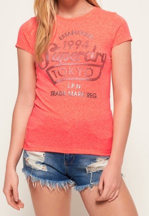 Camiseta de manga corta color coral con un estampado gráfico que dice "Superdry Tokyo" y la fecha de creación. Combinada con pantalones cortos de mezclilla desgastada.